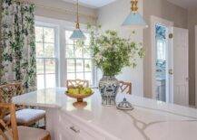 Vintage blue lanterns light a white angled kitchen island topped with a marble-look countertop and paired with tan bamboo counter stools. Windows are covered in white and green botanical curtains.