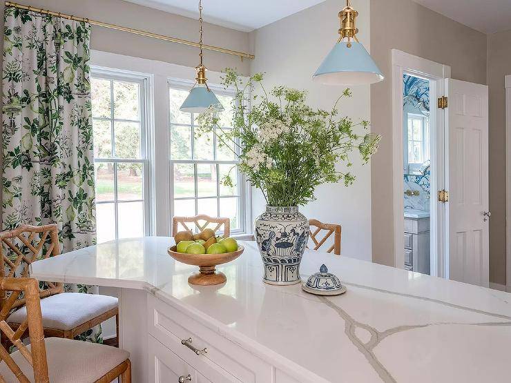Vintage blue lanterns light a white angled kitchen island topped with a marble-look countertop and paired with tan bamboo counter stools. Windows are covered in white and green botanical curtains.