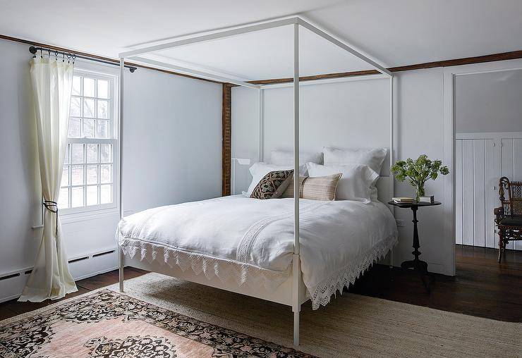 Bedroom features a white canopy bed with white bedding, a pink and beige rug layered atop a tan jute rug and a round black French bedside table.