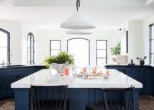A blue wooden center island is matched with black spindle stools placed on a wood floor at a white marble countertop lit by white lanterns.