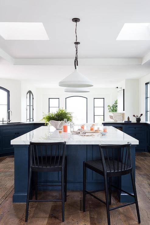 A blue wooden center island is matched with black spindle stools placed on a wood floor at a white marble countertop lit by white lanterns.