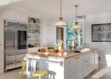Yellow spindle stools sit at a white kitchen island boasting a white towel bar and a butcher block countertop lit by two white and gold lanterns. The island is accented with brass hardware and completed with a sink with an antique brass vintage style faucet.