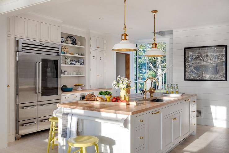 Yellow spindle stools sit at a white kitchen island boasting a white towel bar and a butcher block countertop lit by two white and gold lanterns. The island is accented with brass hardware and completed with a sink with an antique brass vintage style faucet.