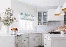A white angled kitchen boasts a white island donning brass pulls and a marble top lit by two Aerin Agnes pendants. A marble waterfall top accents a peninsula.