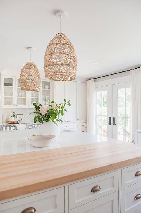 Wicker basket chandeliers illuminate a white kitchen island boasting drawers with brass cup pulls and a maple and quartz countertop.