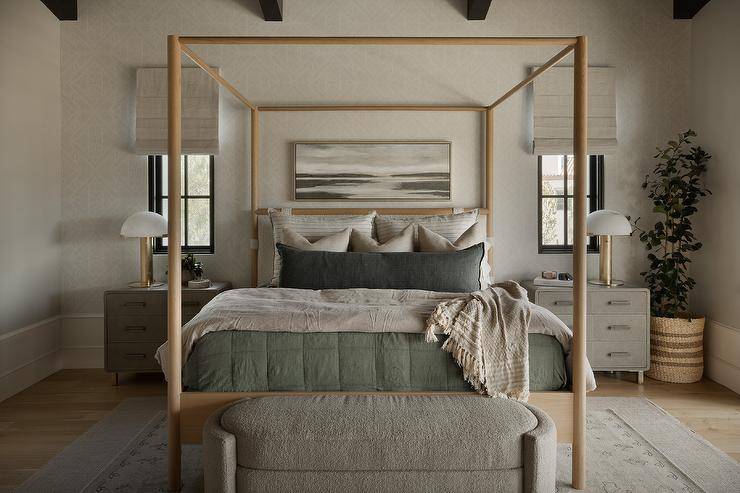 Bedroom features a light brown wooden canopy bed with green and gray bedding flanked by gray nighstands lit by brass and alabaster lamps, an oval gray boucle bench at the foot of the bed and a tall potted plant.