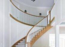 A light stained wood herringbone floor leads to a winding light stained wood staircase accented with a gray diamond pattern runner and white spindles finished with a light stained wood railing.