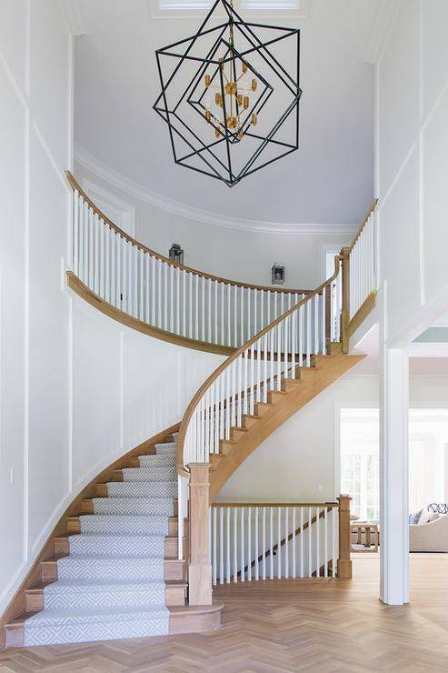 A light stained wood herringbone floor leads to a winding light stained wood staircase accented with a gray diamond pattern runner and white spindles finished with a light stained wood railing.