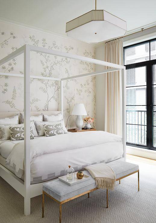white wooden canopy bed, a gold and gray bench at the foot of the bed and a light gray lamp against gray wallpaper accent wall.