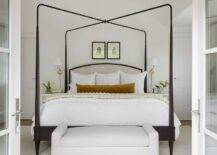 A white settee is positoined at the foot of a gray and black canopy bed draped with an ivory knitted throw blanket and a burnt orange velvet lumbar pillow. Botanical art is hung above the bed, while glass top white nightstands flank the bed and are lit by brass sconces.