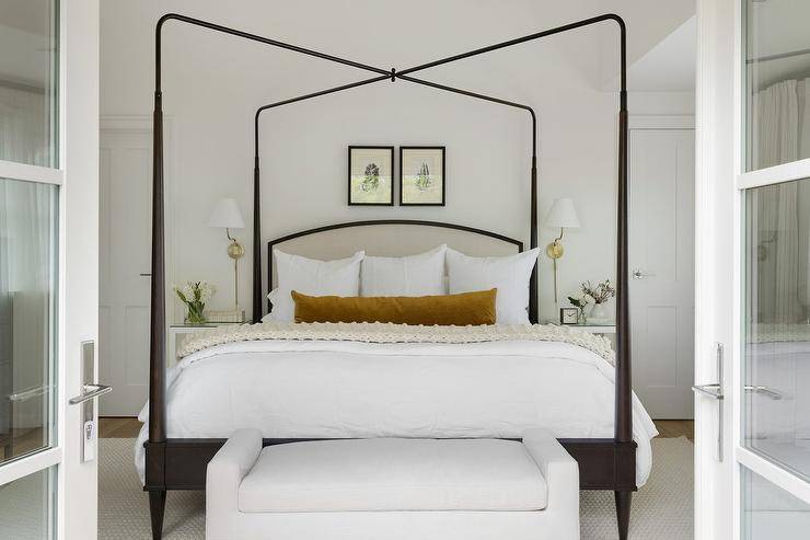 A white settee is positoined at the foot of a gray and black canopy bed draped with an ivory knitted throw blanket and a burnt orange velvet lumbar pillow. Botanical art is hung above the bed, while glass top white nightstands flank the bed and are lit by brass sconces.