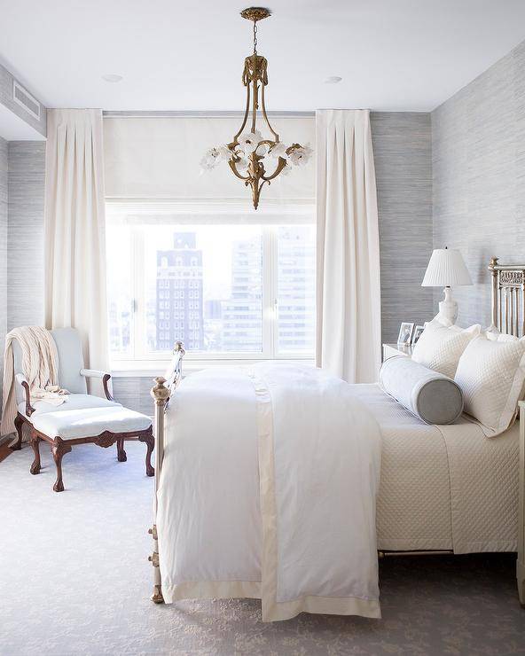 In this romantic white and gray bedroom, ivory curtain cover windows allowing natural light to hit a metal 4 poster bed dressed in white bedding accented with an ivory trim and complemented with a gray bolster pillow. The bed sits on a gray rug in front of a blue-gray grasscloth wallpapered wall lit by an eye-catching chandelier and black and white marble urn lamps.