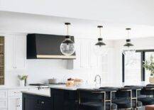 Black velvet stools at a black island with a breakfast bar topped with white quartz in a transitional kitchen. A set of glass and black globe pendant lights align above the island and raised breakfast bar design.