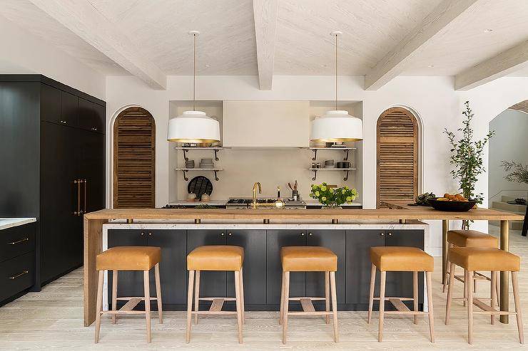 White and gold pendants illuminate an l-shaped live edge breakfast bar finishing a glossy black island donning a marble waterfall countertop. The island is paired with backless brown leather stools.