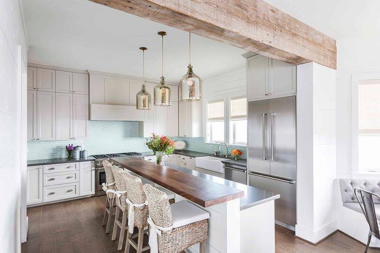 Kitchen features brown wicker stools at a wooden raised island breakfast bar with white shiplap trim, illuminated by mercury glass lanterns and light gray cabinets with blue glass mini subway tiles.