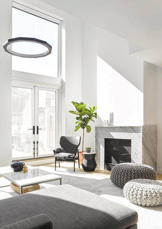 Chic modern living room boasts gray knitted stools placed on a gray rug in front of a modern gray marble fireplace, as a black accent table is paired iwth a black leather chair located in front of a fiddle leaf fig.