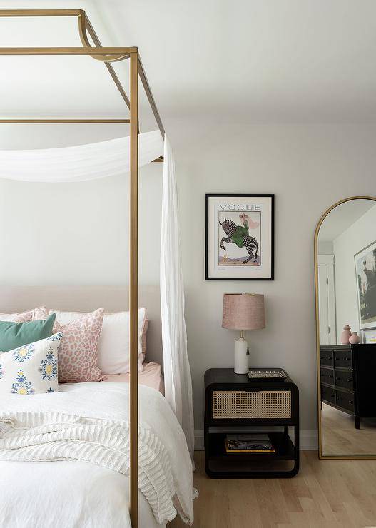 Romantic white curtains complement a gold canopy bed dressed in white and pink bedding complemented with pink animal print pillows and green pillows. The bed is paired with a black cane nightstand lit by an alabaster lamp with a mauve linen lamp shade.