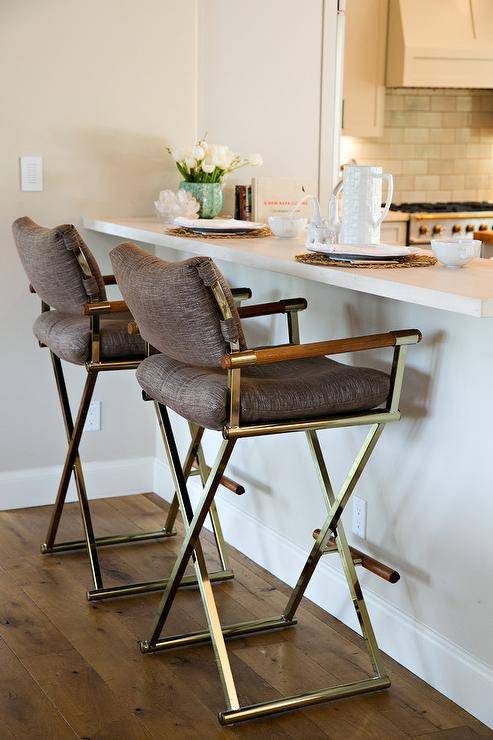 A white kitchen peninsula is paired with a set of vintage brass folding bar stools that showcase comfort with its brown linen cushions. These x base counter stools with a relaxed back support, boast a midcentury modern feel.