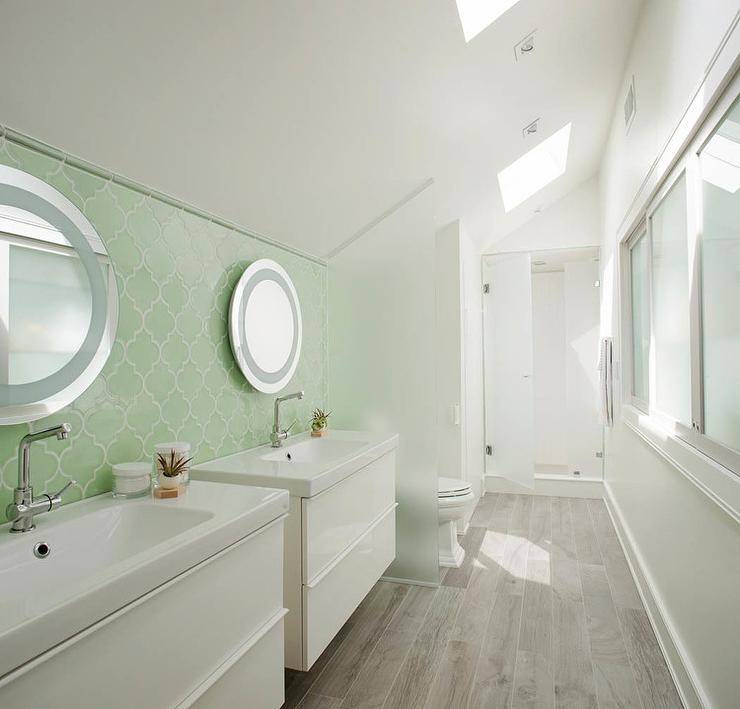 Fabulous galley bathroom features separate side by side floating washstands and round vanity mirrors lining a wall clad in a green Moroccan tile backsplash beneath a sloped ceiling fitted with skylights. A frosted glass partition separates the bathroom vanities from the toilet and walk-in shower.
