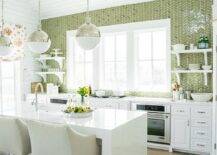 White and green kitchen features white cabinets paired with white quartz countertops and a ceiling height green herringbone tiled backsplash. An integrated gas cooktop stands under windows flanked by stacked white shelves and oven to the left and stacked shelves and oven to the right. Two Hicks Pendants hang over a white quartz waterfall kitchen island fitted with a sink and gooseneck faucet lined with ivory swoop arm bar stools.