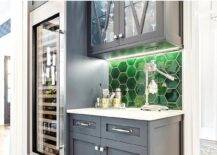 Contemporary home bar is fitted with blue cabinets donning polished nickel hardware and fixed against emerald green glass hexagon backsplash tiles under glass front mullion cabinets and beside a full size glass front wine fridge.