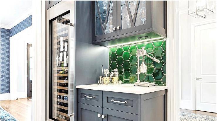 Contemporary home bar is fitted with blue cabinets donning polished nickel hardware and fixed against emerald green glass hexagon backsplash tiles under glass front mullion cabinets and beside a full size glass front wine fridge.