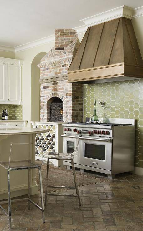 A green hex tile backsplash surrounding stone accents keeps with the rustic theme in this kitchen. Green honeycomb tiles with white grout frame a stainless steel Wolf dual range next to a red brick pizza oven over white built-in wine storage. Over the range, a gold French kitchen hood also acts as the room's focal point complimenting brick herringbone kitchen floors.