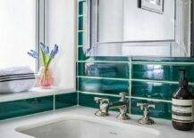 A blue floating washtsand with a polished nickel cross handle faucet is mounted against stacked emerald green tiles under a framed inset medicine cabinet.