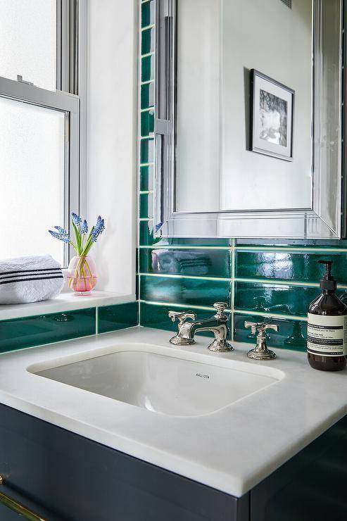 A blue floating washtsand with a polished nickel cross handle faucet is mounted against stacked emerald green tiles under a framed inset medicine cabinet.