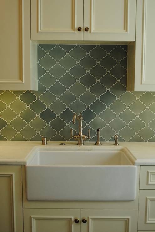 Stunning kitchen with hook spot faucet over farm sink framed by beaded cream cabinets accented with brass hardware and sleek white countertops below a green arabesque tiled backsplash.