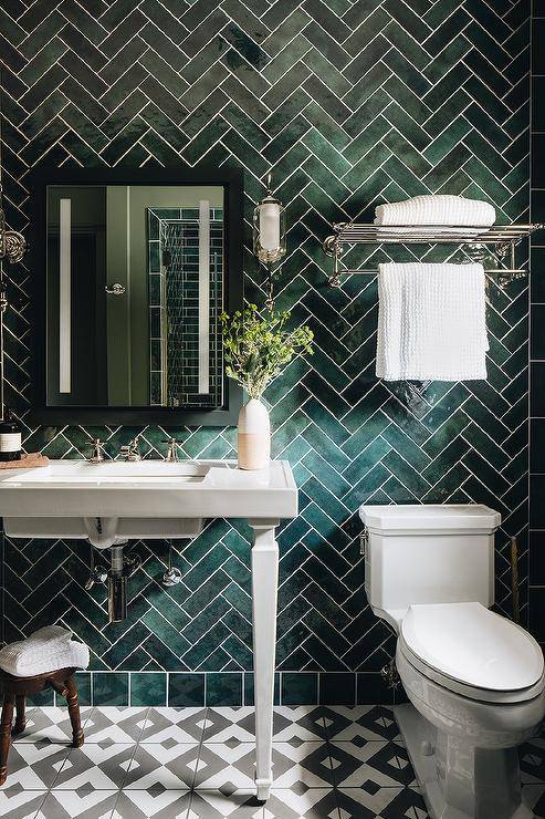 Bathroom features a white pedestal sink on a green herringbone pattern tiled wall, a black framed medicine cabinet, a nickel train towel rack over the toilet and white and gray diamond floor tiles.