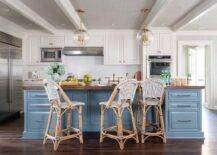 Well appointed white and blue kitchen boasts Serena & Lily Riviera Counter Stools placed at a blue center island accented with a walnut butcher block countertop lit by brass and glass globe lanterns hung from a gray wallpapered ceiling.