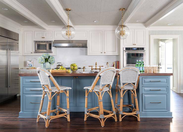 Well appointed white and blue kitchen boasts Serena & Lily Riviera Counter Stools placed at a blue center island accented with a walnut butcher block countertop lit by brass and glass globe lanterns hung from a gray wallpapered ceiling.