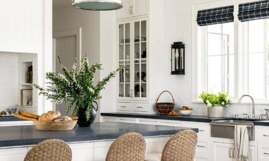 Kitchen features a white beadboard ceiling with white industrial lighsts that illuminate a honed black marble top island with tan woven stools, white cabinets with nickel pulls and honed black marble countertop, a French sconce on glazed white tiles, a vintage runner and blue plaid roman shades over a stainless steel apron sink with towel bar.