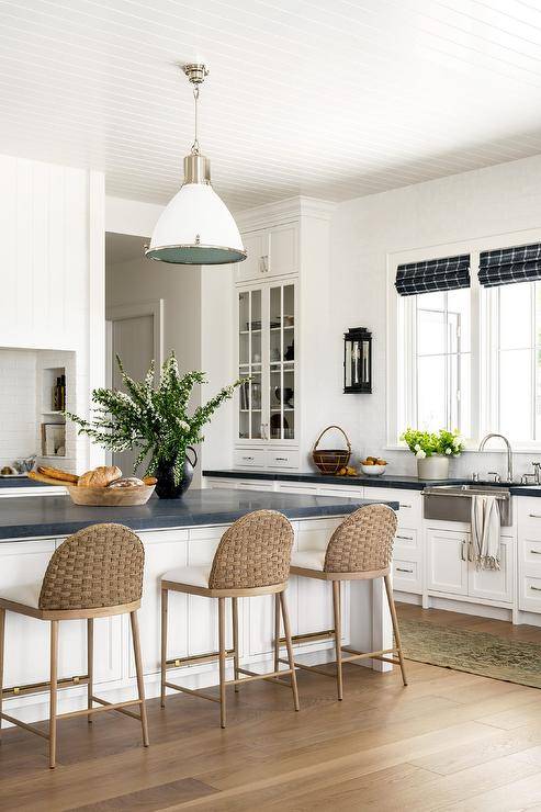 Kitchen features a white beadboard ceiling with white industrial lighsts that illuminate a honed black marble top island with tan woven stools, white cabinets with nickel pulls and honed black marble countertop, a French sconce on glazed white tiles, a vintage runner and blue plaid roman shades over a stainless steel apron sink with towel bar.