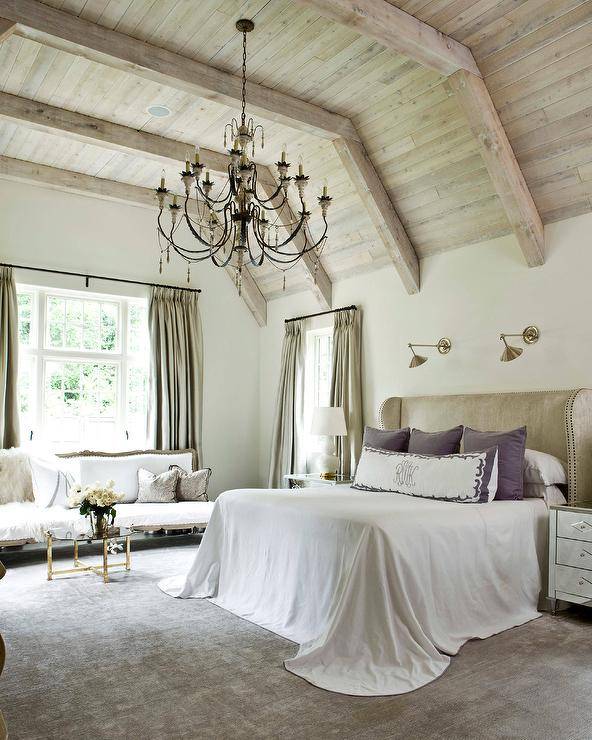 From a stunning plank vaulted ceiling accented with stunning rustic wood beams, a French candle chandelier hangs in front of a taupe leather wingback headboard lit by two brass wall sconces and positioned behind a bed dressed in white bedding topped with purple velvet pillows accented with a white and purple monogrammed lumbar pillow.The bed is flanked by mirrored nightstands lit by white double gourd lamps and sits beside a window dressed in taupe silk French pleat curtains. In front of a window an adjacent wall, a long white French settee is placed behind a round glass and brass cocktail table.