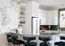 A curved granite top peninsula seats teal t-back leather stools, while white display shelves are mounted to the side of a recessed stainless steel refrigerator located beneath white cabinet with brass knobs.