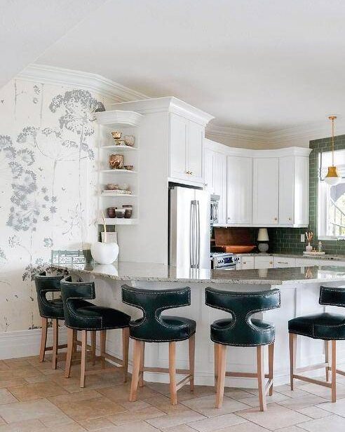 A curved granite top peninsula seats teal t-back leather stools, while white display shelves are mounted to the side of a recessed stainless steel refrigerator located beneath white cabinet with brass knobs.