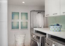 Gorgeous laundry room with gray front loading washer and dryer below white folding counter topped with ceiling height cabinets over a mint green glass brick mosaic tiled backsplash. Mint green framed art hangs over the laundry hamper at the back of the space beside a chrome drying rail over light stone tiled floors.