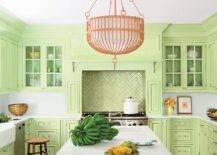 A coral pink chandelier hangs over a light green island topped with white Corian countertops fitted with a sink and a polished nickel hook and spout faucet. Behind the island a conceal light green hood is fixed above green metallic herringbone cooktop tiles accenting a stainless steel oven range. The hood is flanked by glass front upper cabinets mounted above light green lower cabinets lined with a white corian backsplash and finished with a stainless steel dishwasher and a farmhouse sink.