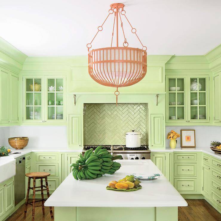 A coral pink chandelier hangs over a light green island topped with white Corian countertops fitted with a sink and a polished nickel hook and spout faucet. Behind the island a conceal light green hood is fixed above green metallic herringbone cooktop tiles accenting a stainless steel oven range. The hood is flanked by glass front upper cabinets mounted above light green lower cabinets lined with a white corian backsplash and finished with a stainless steel dishwasher and a farmhouse sink.