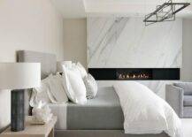 Gray modern bedroom boasts a modern gray bed accented with white and gray bedding and flanked by tan wood nightstands topped with black lamps. The room is warmed by a lava rock fireplace fixed facing gray velvet accent chairs