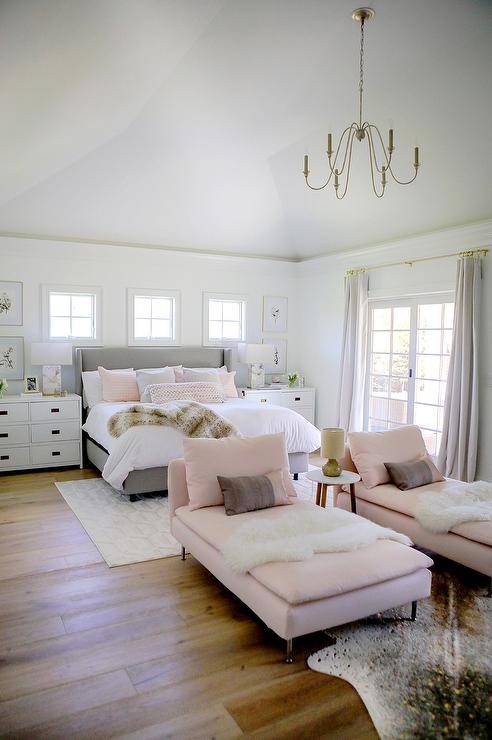 Stylish pink and gray contemporary bedroom boasts a lounge area featuring pink chaise lounges topped with white sheepskin throws and pink and brown pillows. The chaise lounges sit on a metallic cowhide rug beneath a brass chandelier.
