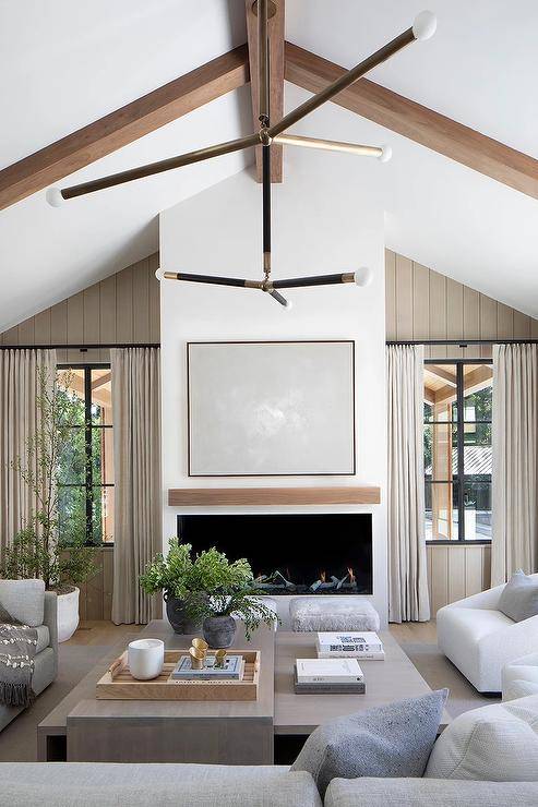 A light gray abstract art piece hangs over a modern fireplace fitted with a wood floating mantel and flanked by windows framed by taupe plank trim and covered in beige curtains.