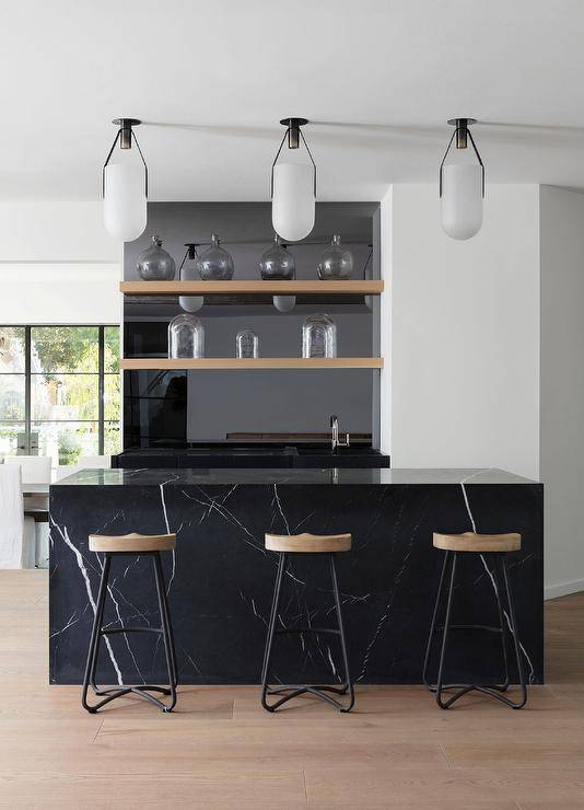 Modern kitchen features a honed black marble bar with wooden shelves, polished black marble countertop and wood and metal tractor stools lit by white pendants.