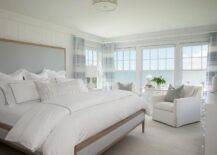 Brown and blue linen bed with white hotel bedding accented with blue borders in a cottage ocean view master bedroom. White linen skirted accent chairs join a white side table at the bedroom windows boasting a stunning view of a serene ocean and clear skies.