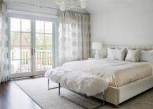 Romantic cream and gray bedroom boasts light gray walls and a cream suede bed accented with gold sheets and gray silk pillows. In front of the bed, a sheepskin bench sits on a gray bound sisal rug, while windows are covered in cream and gray dots curtains.