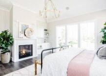 Master bedroom features a cream capiz mirror over a white beveled fireplace mantel, black bed with pink and white bedding, dark wooden floors, lit by a chandelier.