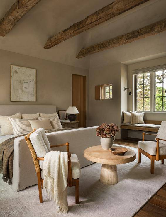Restful cottage bedroom boasts rustic wood ceiling beams mounted to a gray vaulted ceiling over a natural linen slipcovered bed complemented with beige striped pillows. The bed is positioned against a gray wall under a rustic art piece. A round wooden table is flanked by brown wood chairs placed at the foot of the bed on a gray rug.