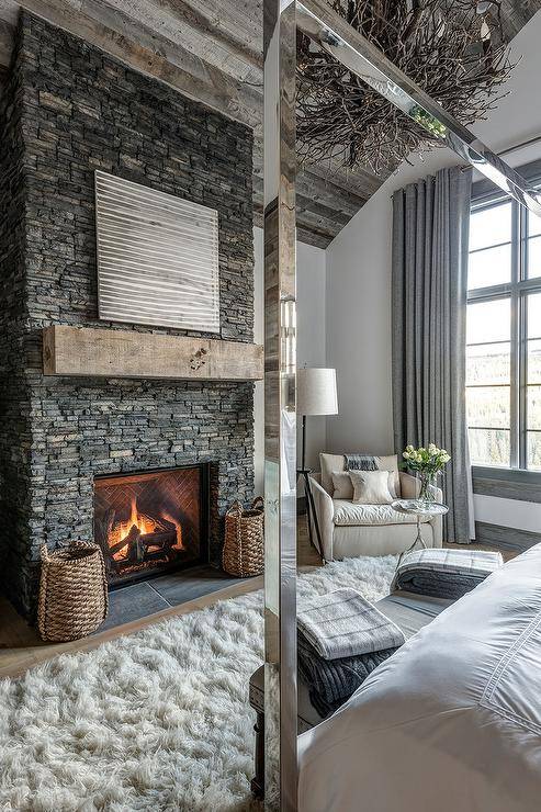 Re،l country ski chalet bedroom is equipped with a gorgeous gray stone fireplace finished with a rustic wood beam mantel fixed below a gray and white abstract art piece mounted under a vaulted wood plank ceiling. Two woven baskets flank the fireplace as a natural linen slipcovered corner chair sits beside a silver leaf faux bois accent table facing a gray bench placed on a white sheepskin rug in front of a chrome canopy bed dressed in white ،tel bedding.
